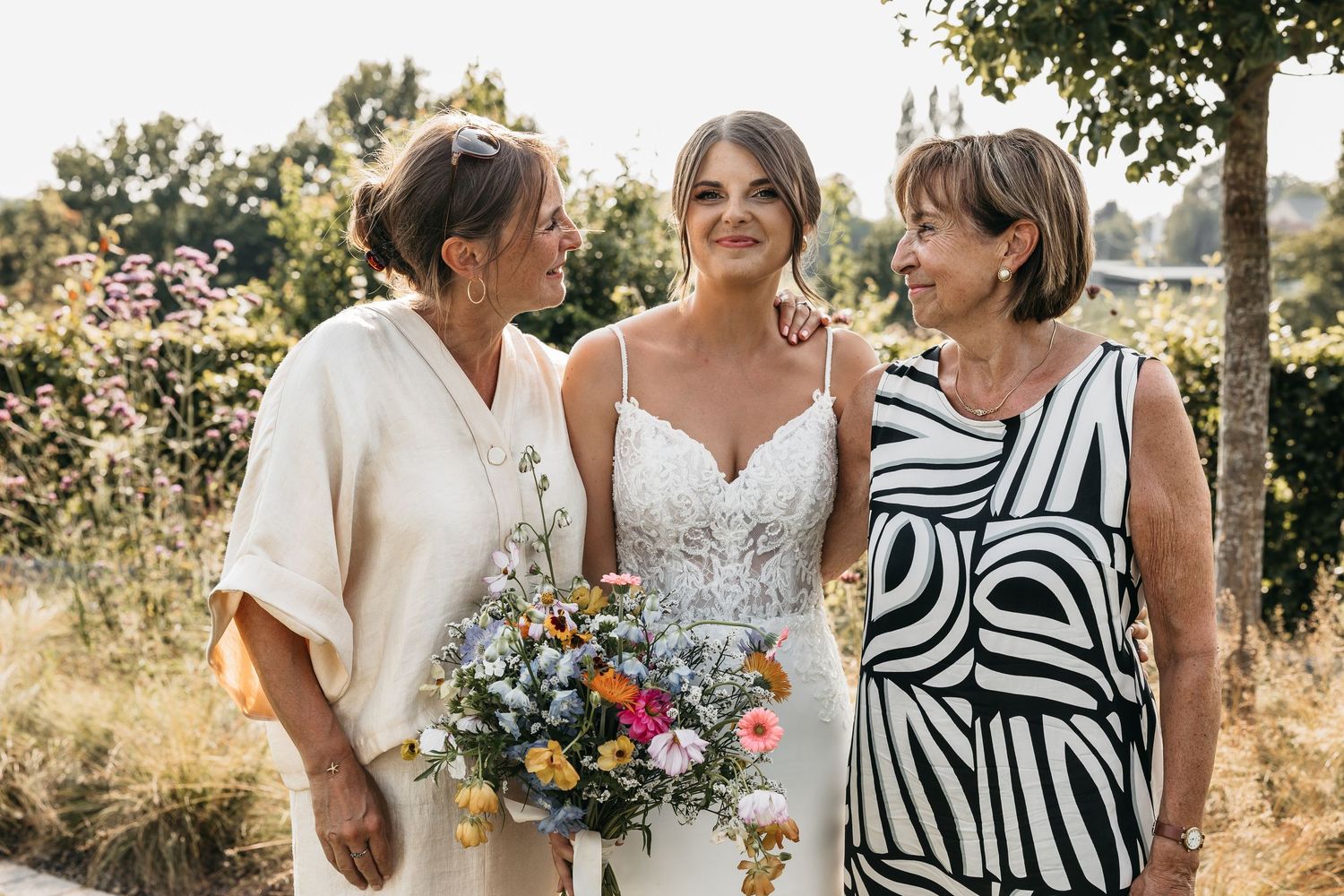 Bride in white lace gown holding wildflower bouquet stands between two women in an outdoor garden setting.