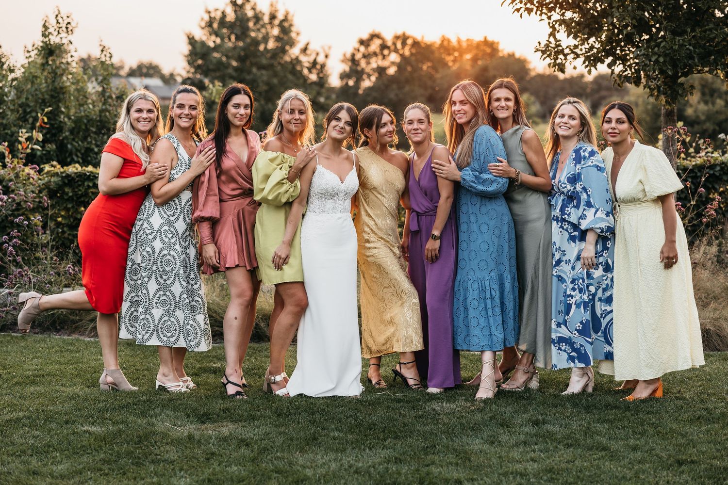 Bride posing with group of female wedding guests in colorful dresses at golden hour.