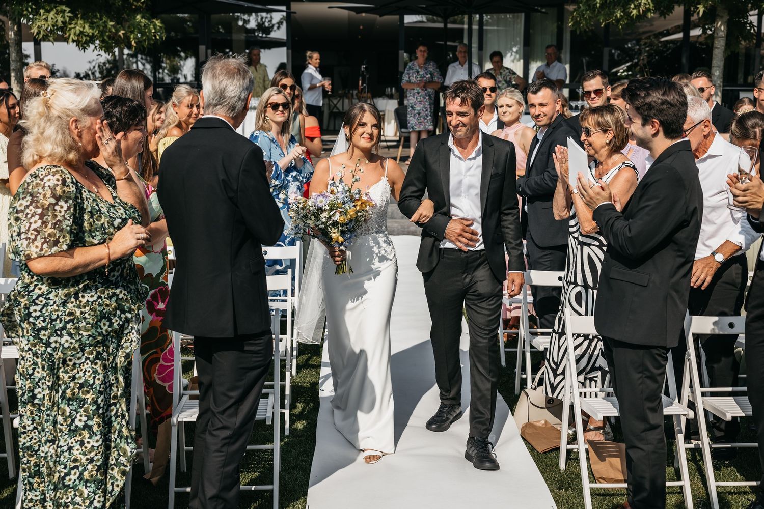 Newlywed couple walks down white aisle after wedding ceremony as guests applaud in celebration.