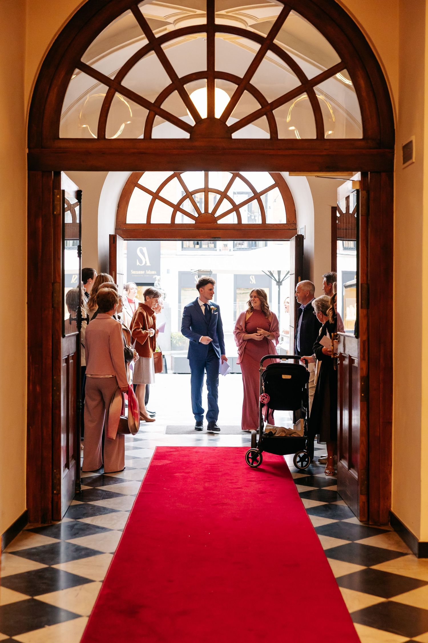 Wedding guests gathering at ornate building entrance with red carpet, groom in blue suit greeting arrivals.