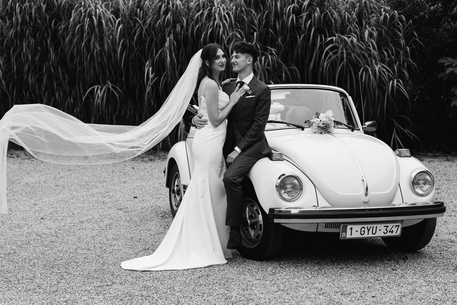 Bride and groom posing in black and white beside a classic Volkswagen Beetle wedding car.