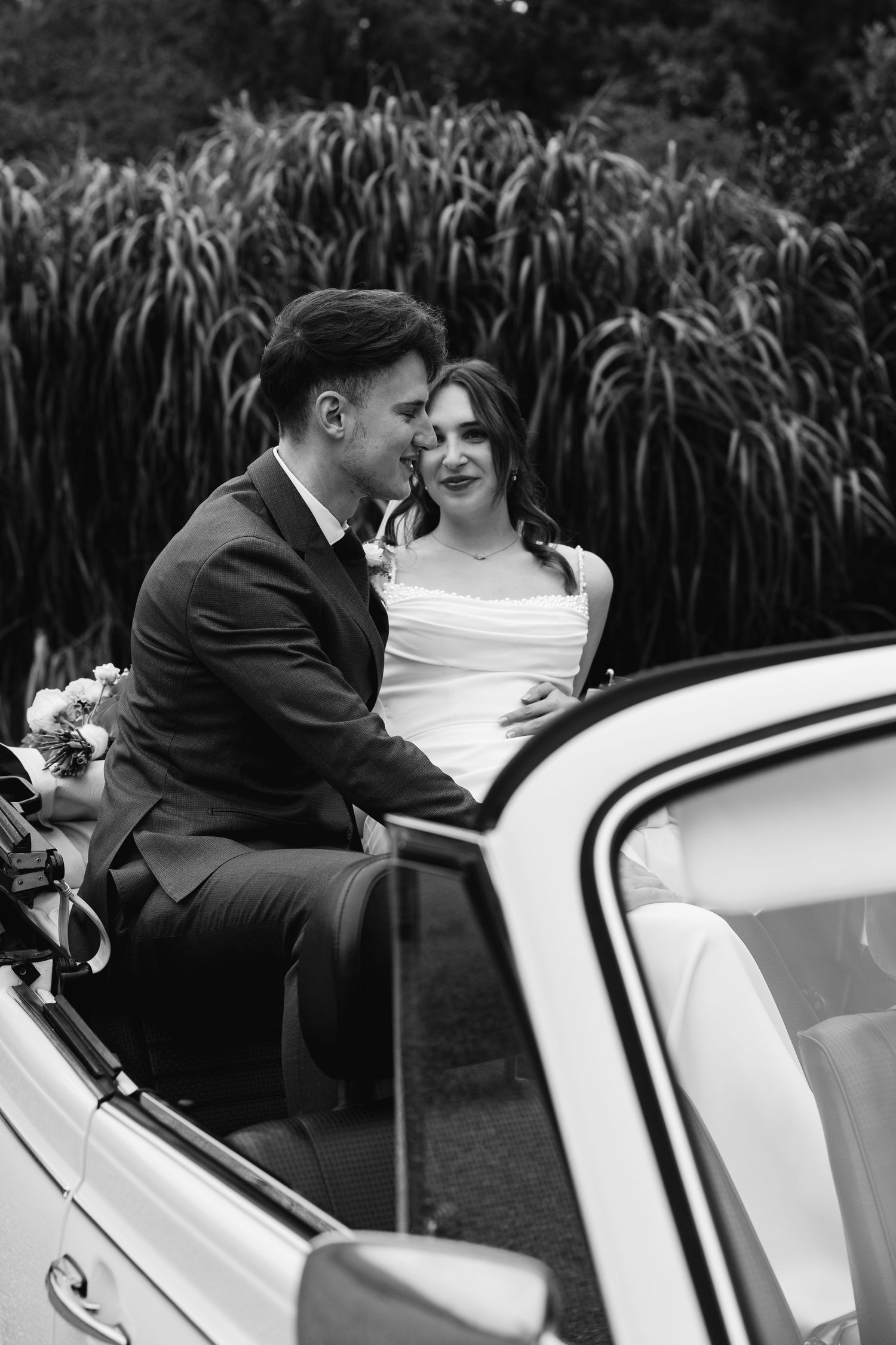 Black and white photo of bride and groom sharing an intimate moment seated in a vintage white convertible car.