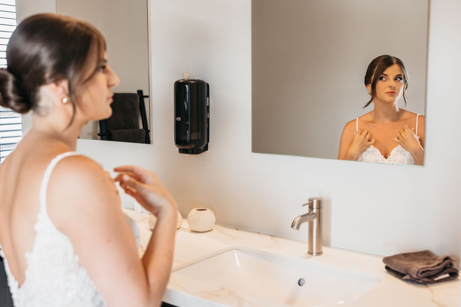 Bride in white wedding dress checking her appearance in bathroom mirror while getting ready.