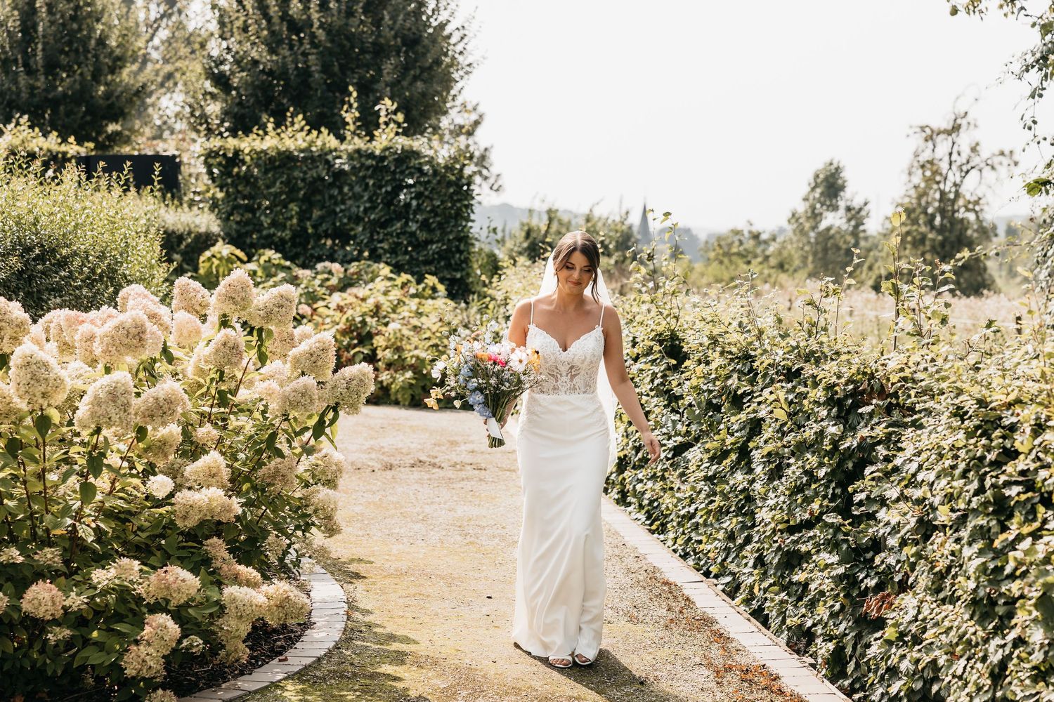 Elegant bride in white lace gown carrying a colorful bouquet walking through a garden.