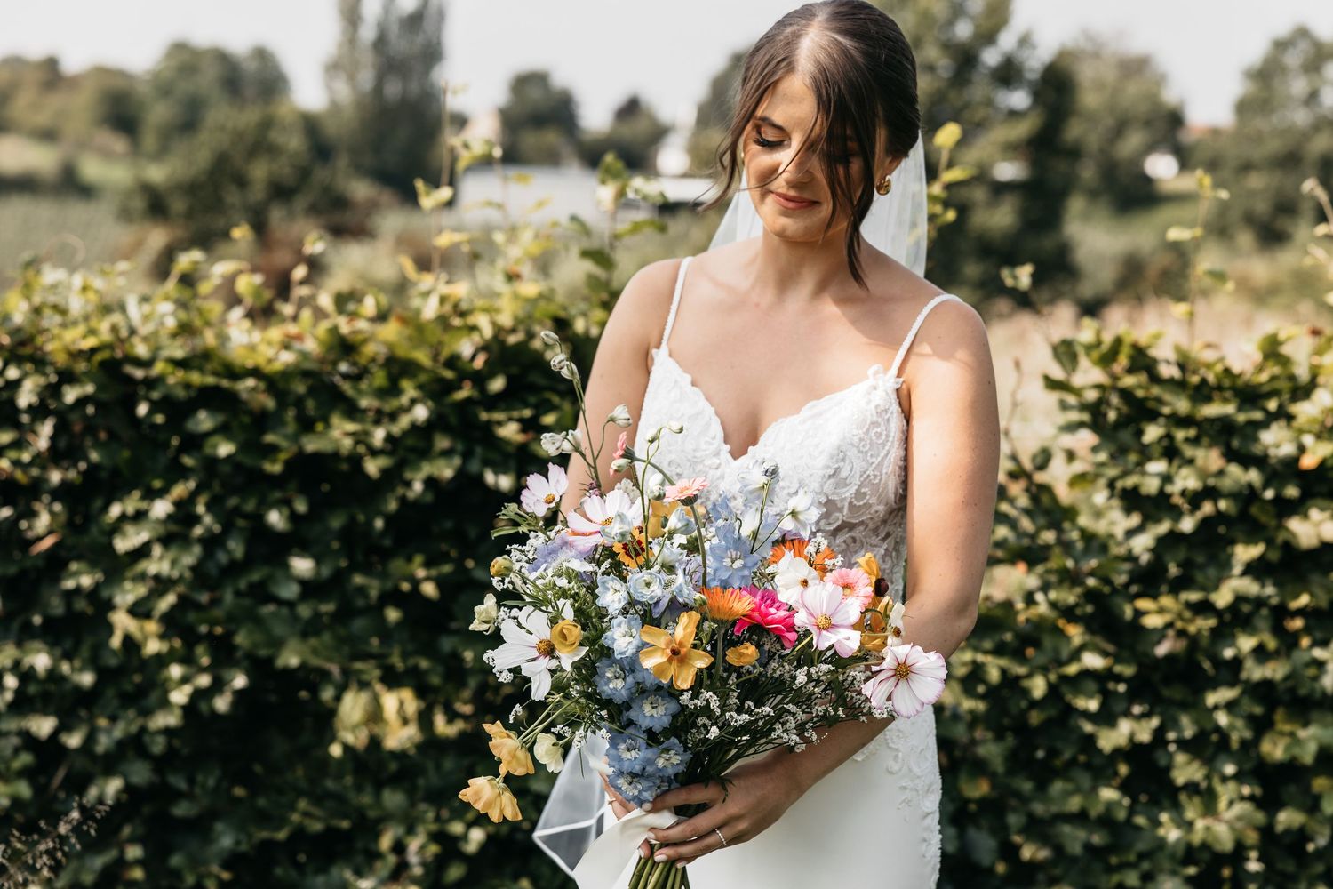 Bride in white lace gown holding colorful wildflower bouquet in sunny garden.