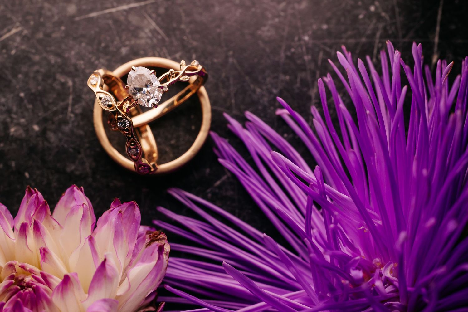 Gold diamond engagement ring and wedding band displayed with purple and pink flowers on dark marble.