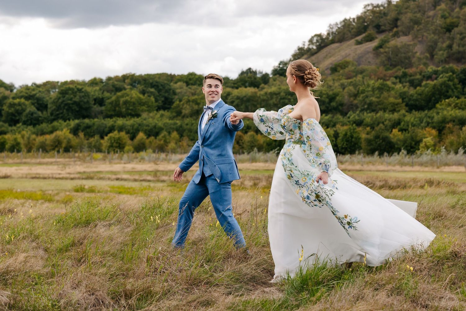 Bride in floral wedding dress and groom in blue suit holding hands while walking through an open field.