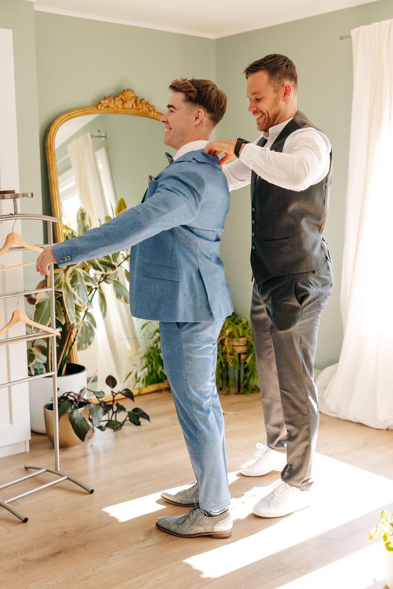 Groomsman adjusting groom's blue suit jacket as both men laugh during preparations.