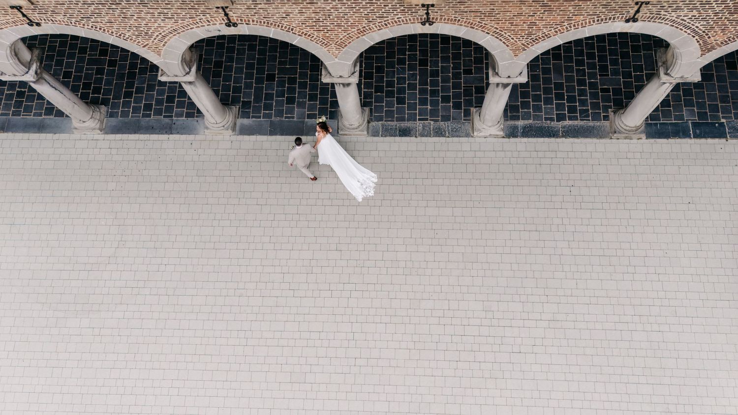 Aerial view of bride and groom embracing on cobblestone plaza beneath arched brick facade.