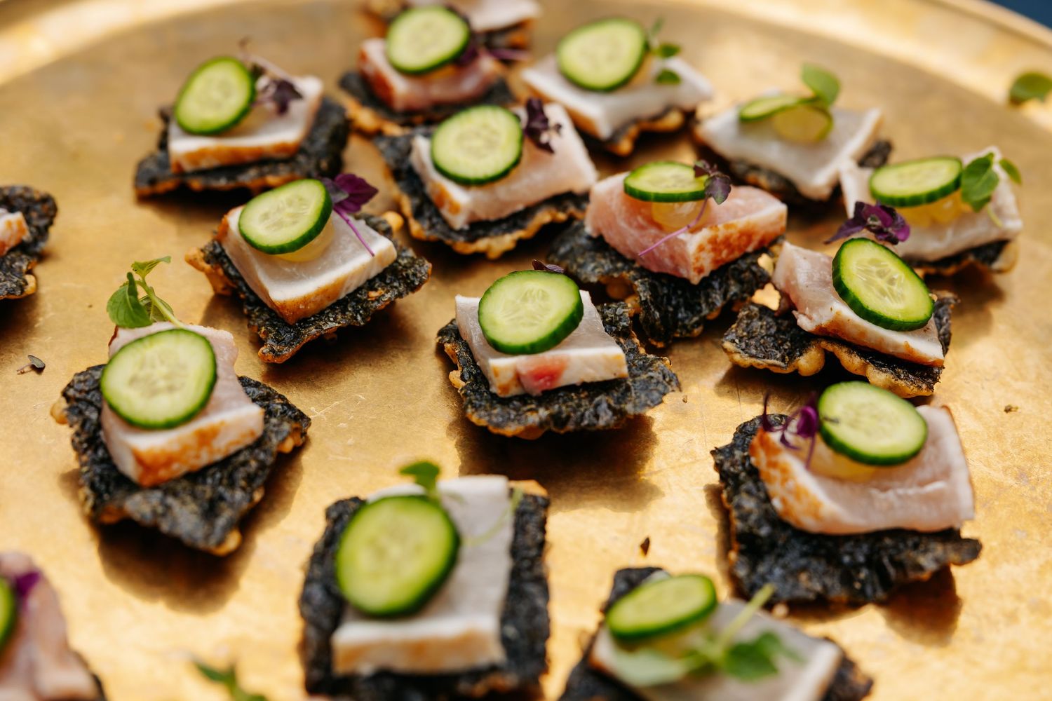 Elegant canapes with tuna, cucumber slices on black crackers served on gold platter.