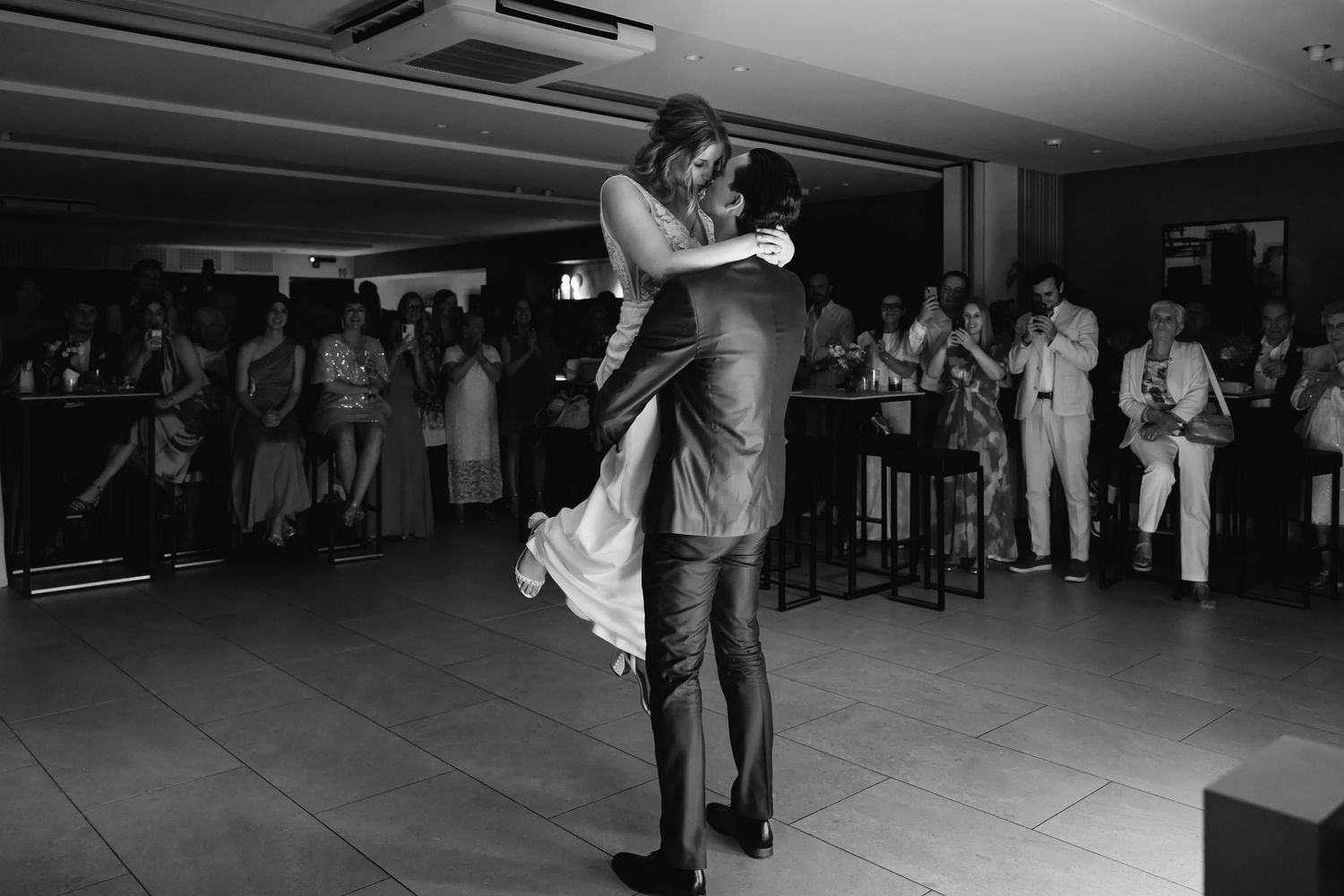 Black and white photo of groom lifting bride during romantic first dance at wedding reception.