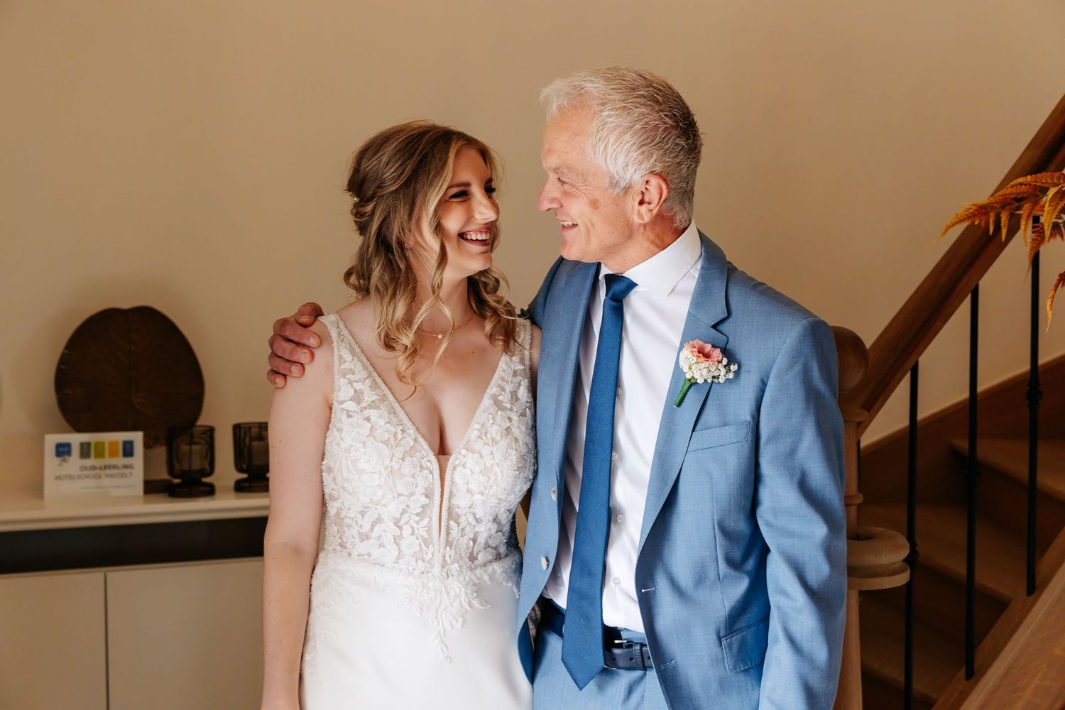 Bride in lace gown and older man in blue suit share a joyful smile near staircase.