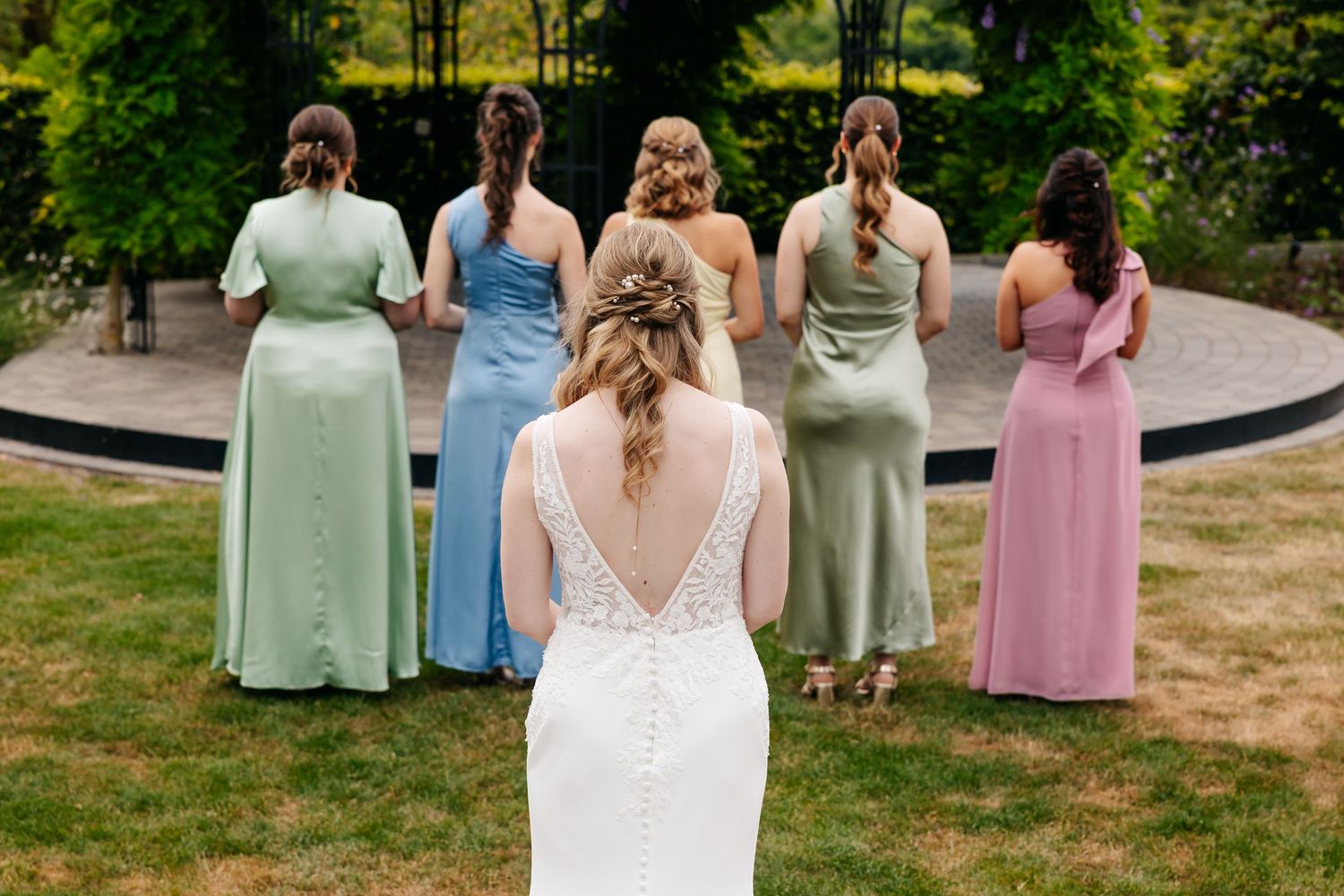 Bride in lace gown stands behind five bridesmaids in pastel dresses facing garden gazebo.