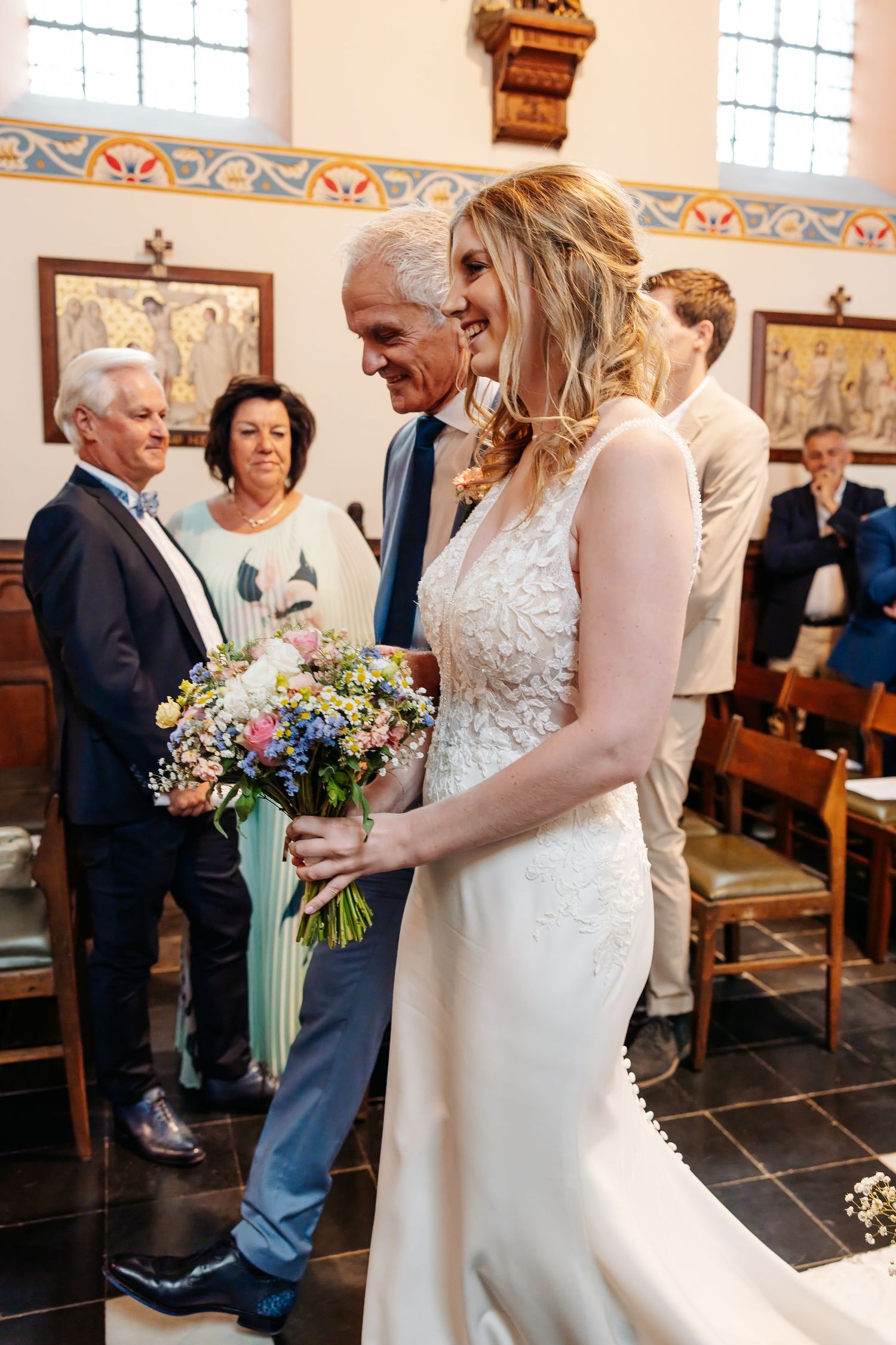 Smiling bride holds wildflower bouquet while walking down church aisle with escort.