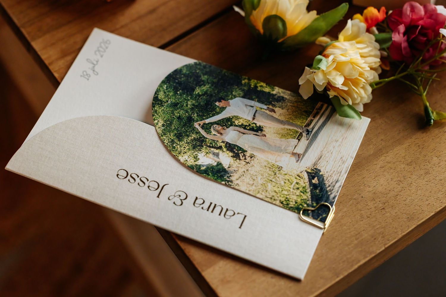 Wedding invitation card for Laura and Jesse with photo and colorful floral bouquet on wooden surface.