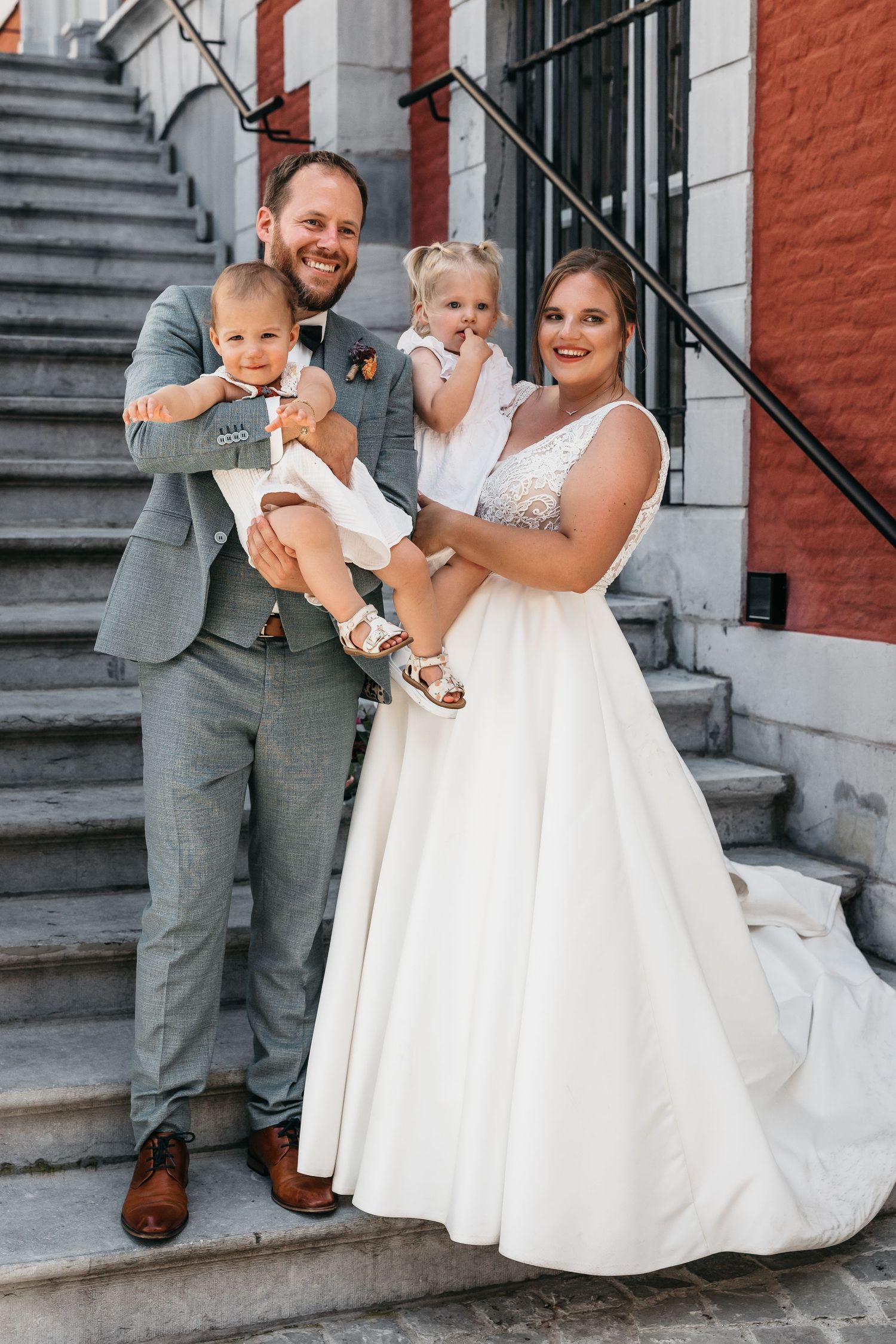 Happy bride and groom posing on steps holding two young children dressed in formal wedding attire.