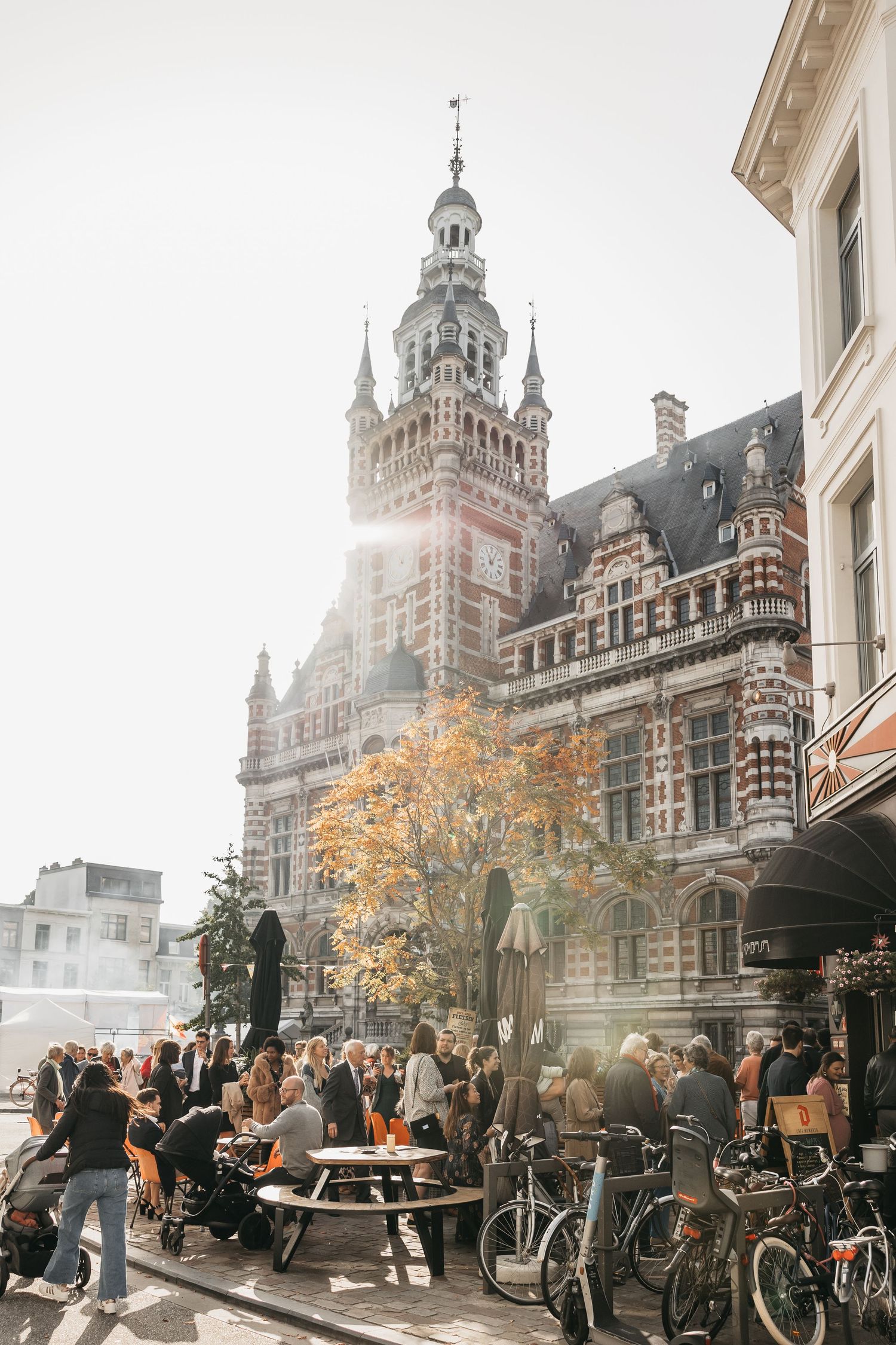 Crowd of people gathered outside historic ornate Belgian building with tower on sunny autumn day.