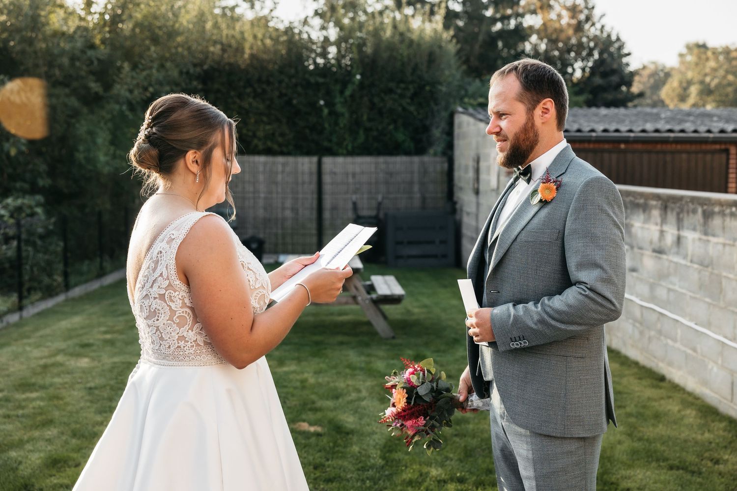 Bride and groom exchange private vows outdoors in garden setting, each holding handwritten vow cards at sunset.