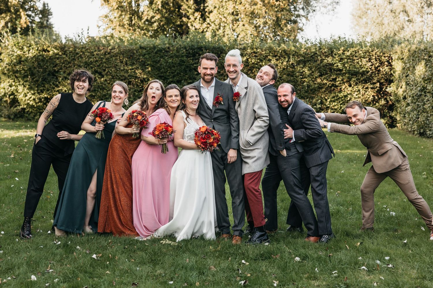 Wedding party group photo on grass with bride, groom, and guests laughing.