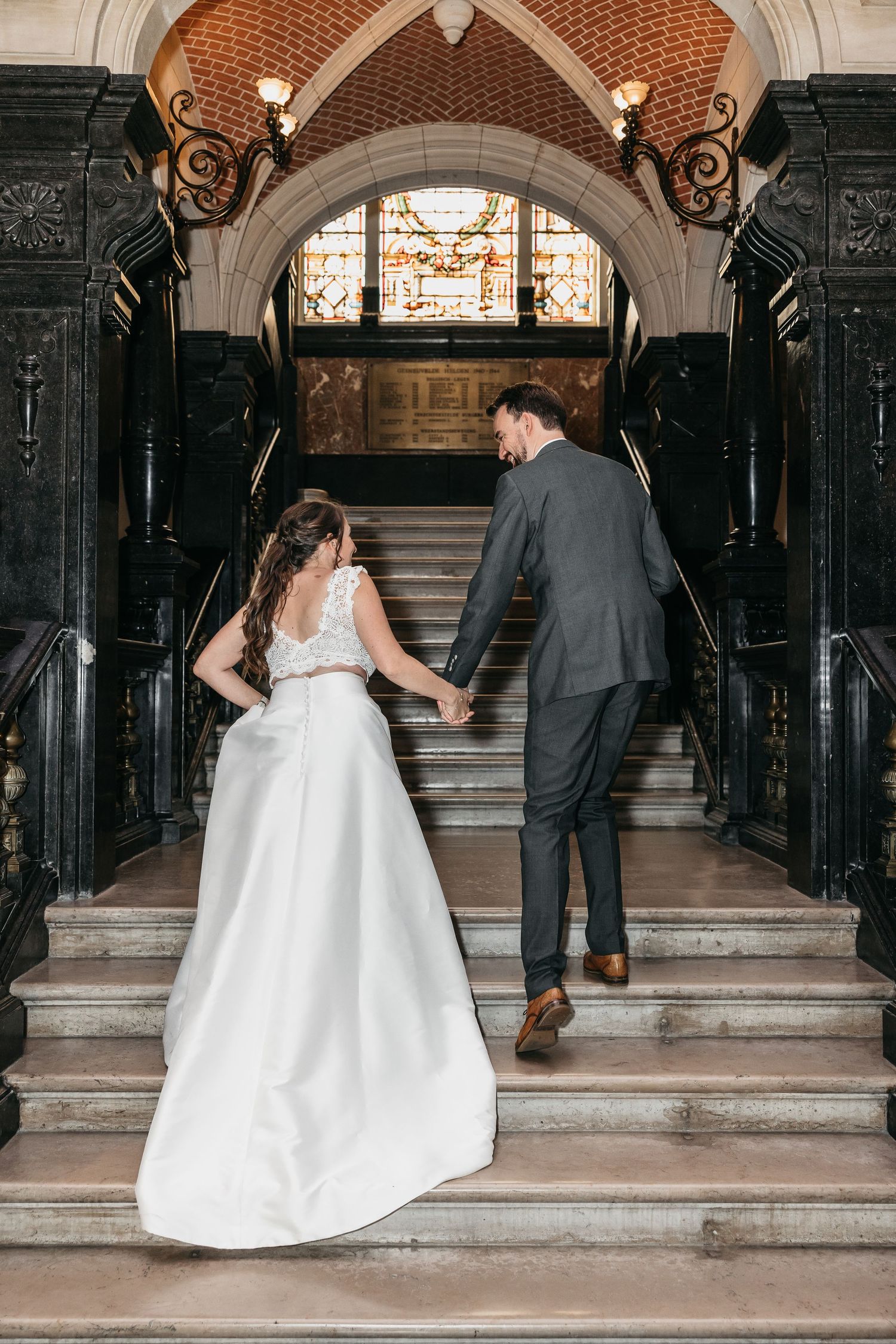 Bride and groom hold hands ascending grand marble staircase beneath stained glass window in historic venue.