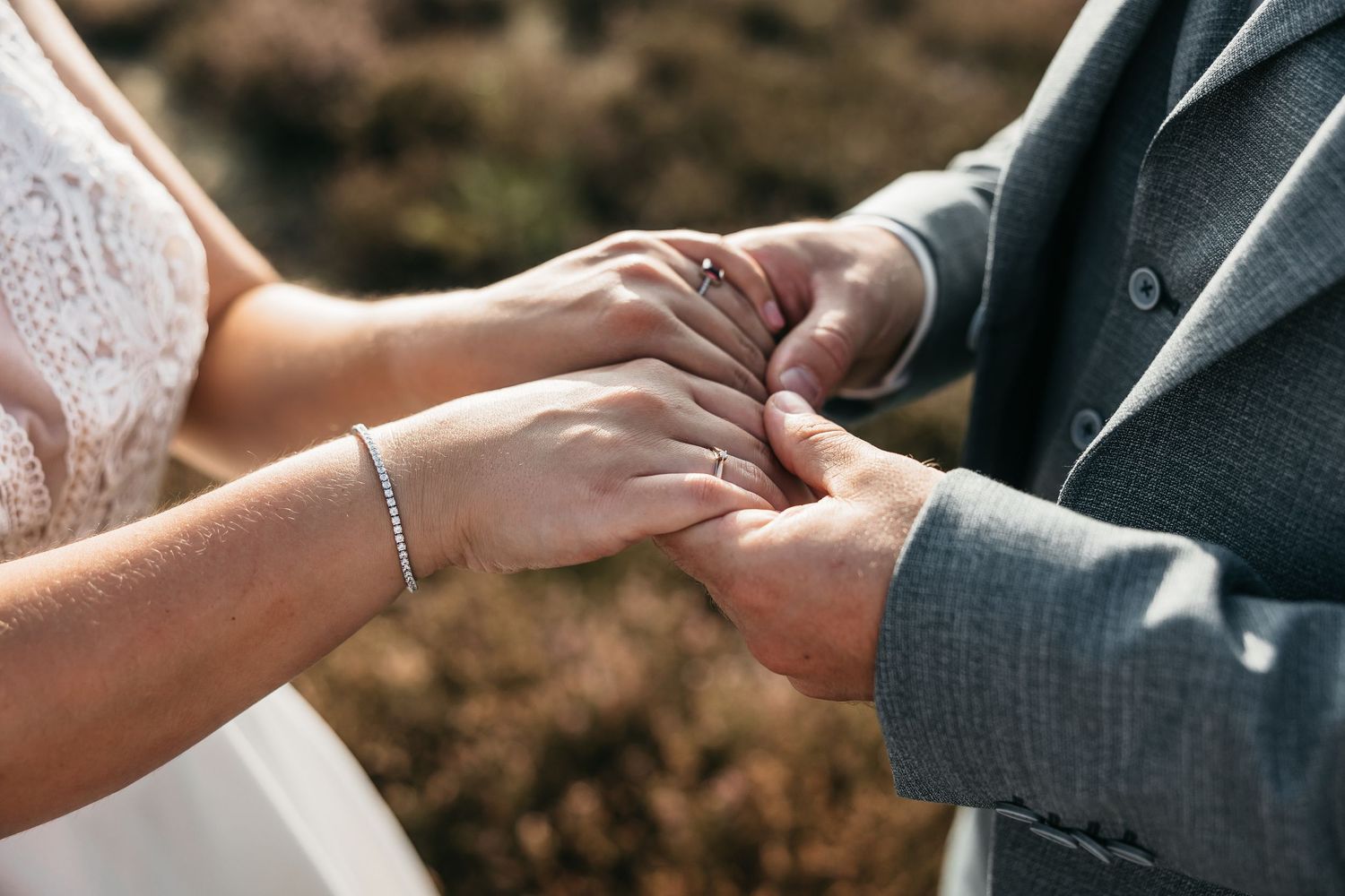Close-up of bride and groom holding hands, showcasing wedding rings and delicate bracelet outdoors.