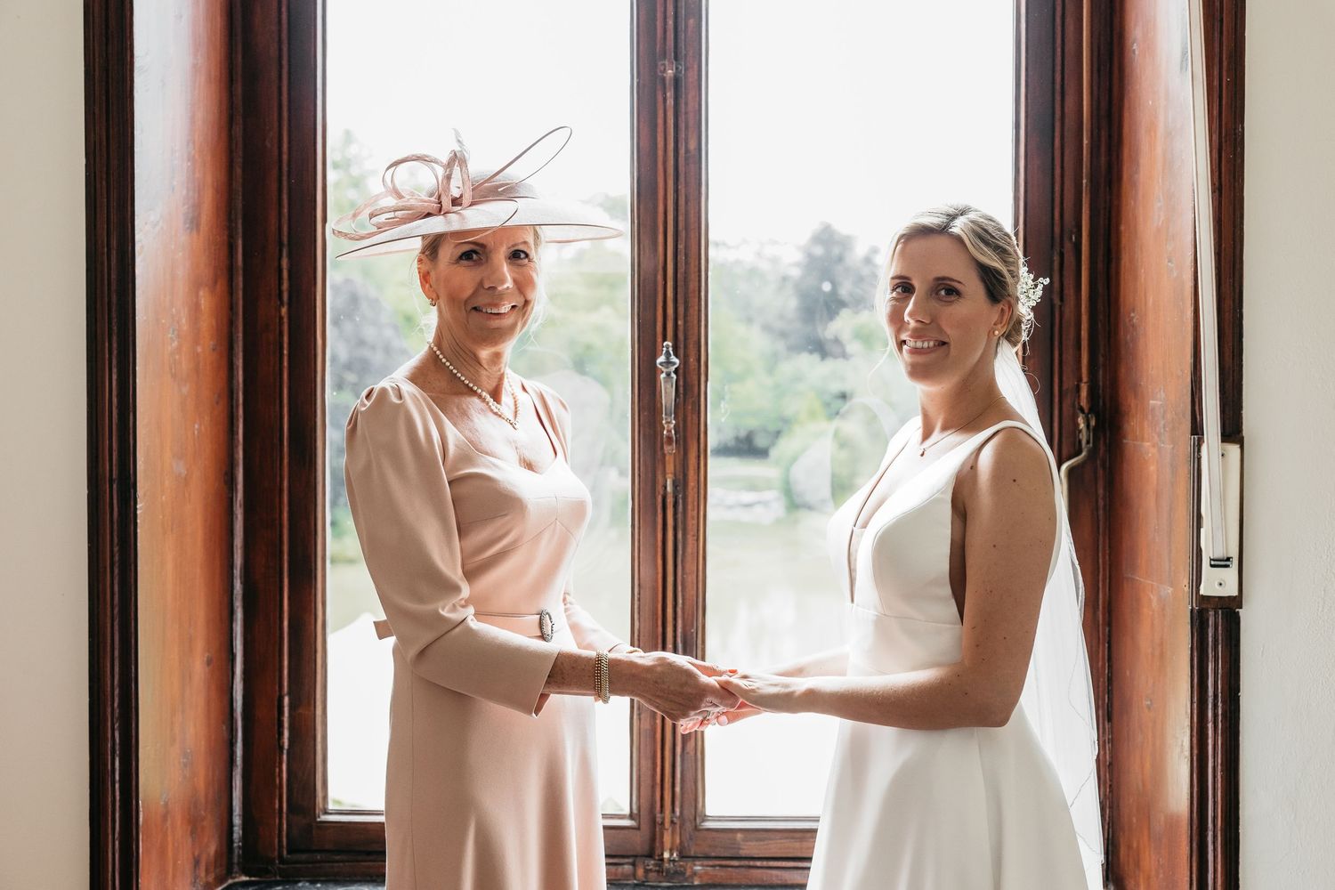 Bride and elegant woman in pink dress holding hands smiling by wooden French doors.