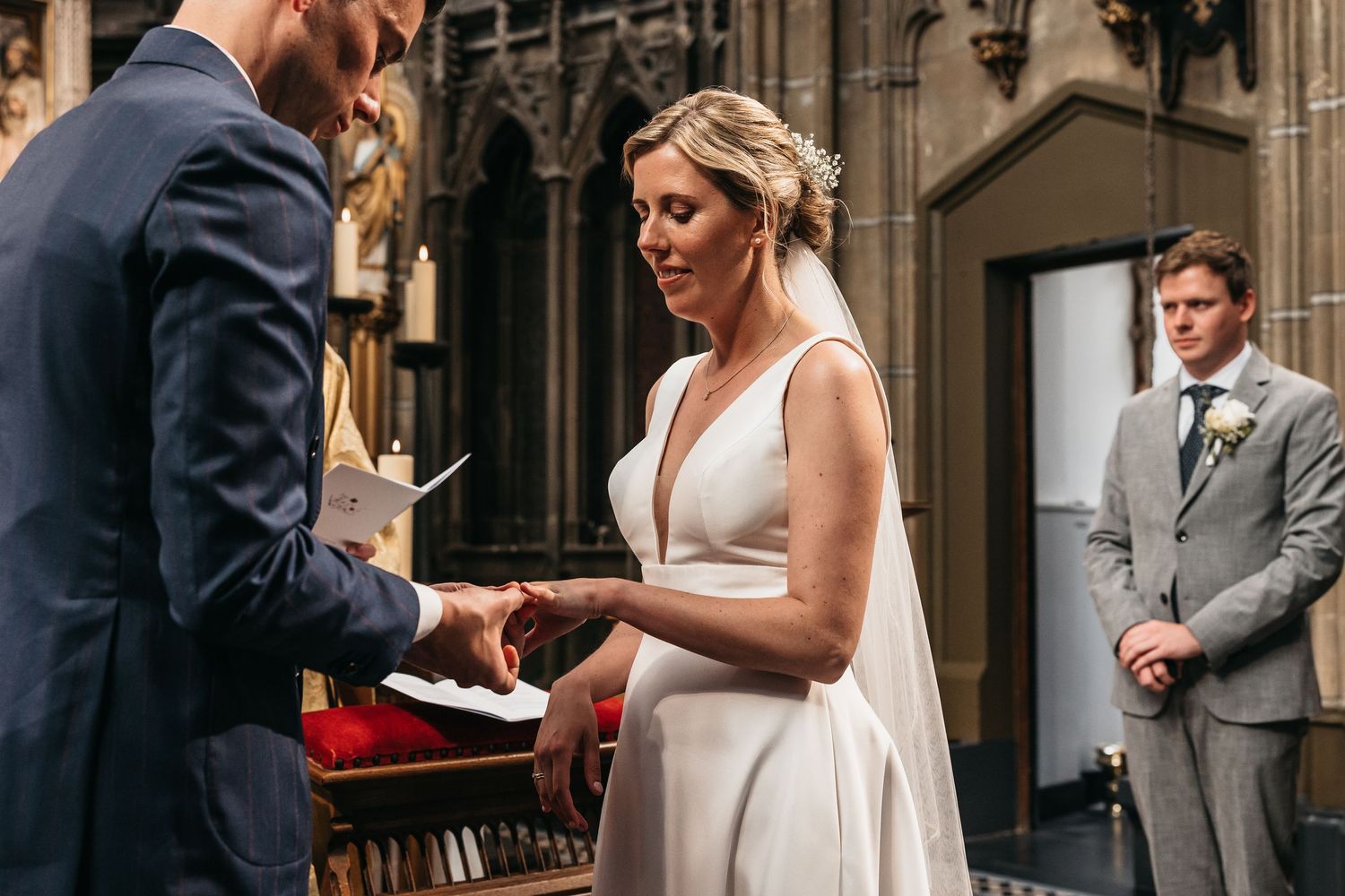 Bride in white gown reads vows while exchanging rings in church.