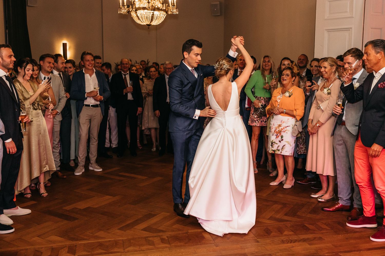 Couple sharing intimate first dance as wedding guests cheer in chandelier-lit ballroom.