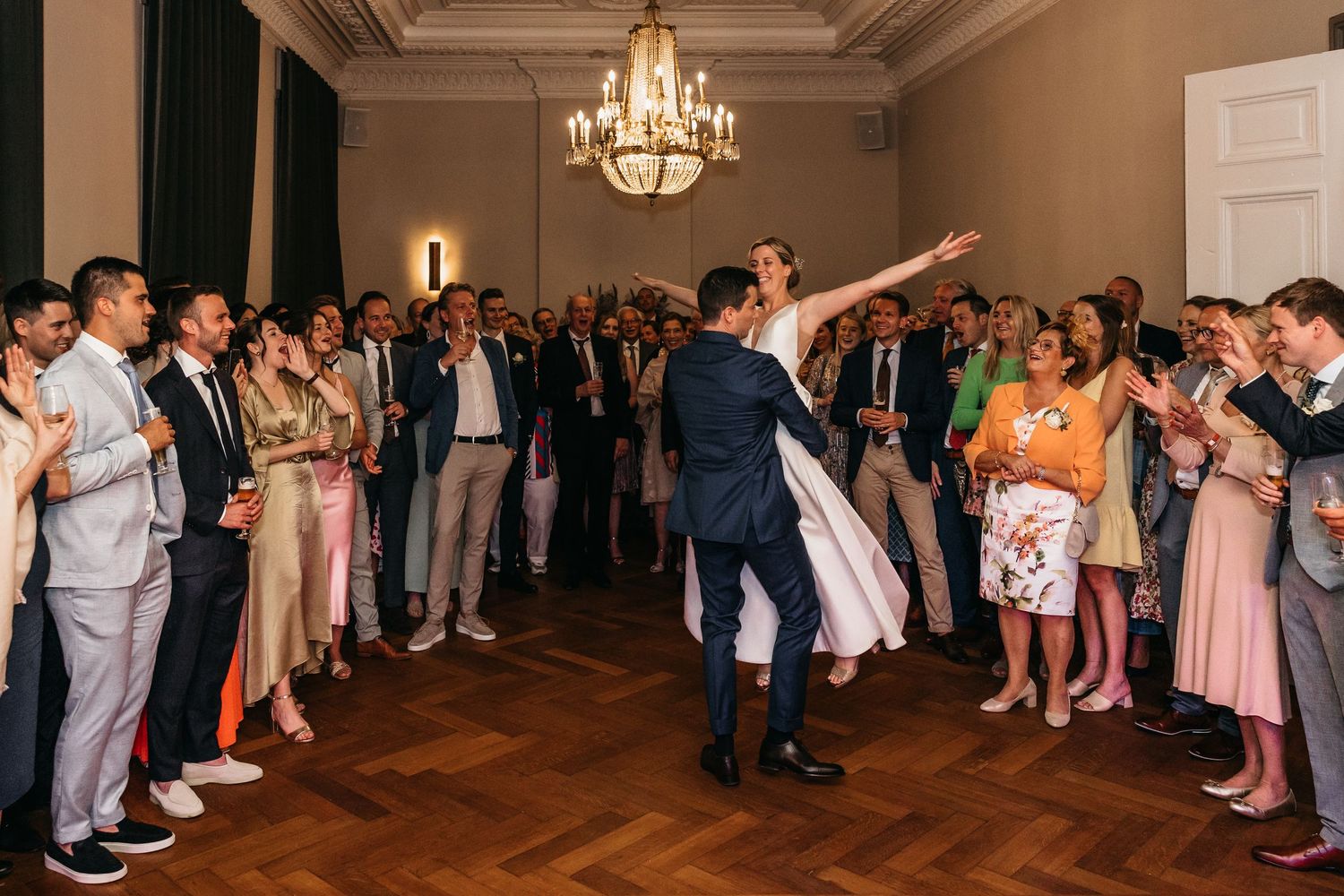Bride and groom share first dance as wedding guests clap and celebrate in elegant chandelier-lit ballroom.