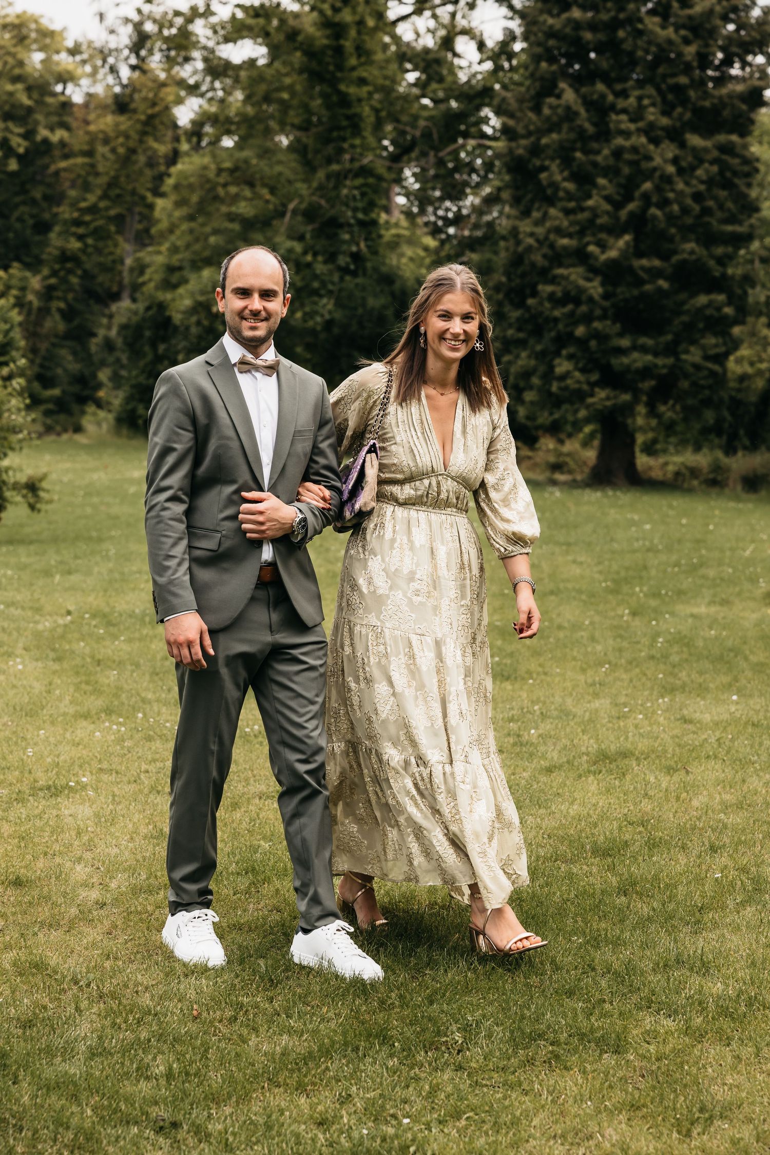 Couple walking on green grass at outdoor wedding, man in gray suit and woman in gold maxi dress.