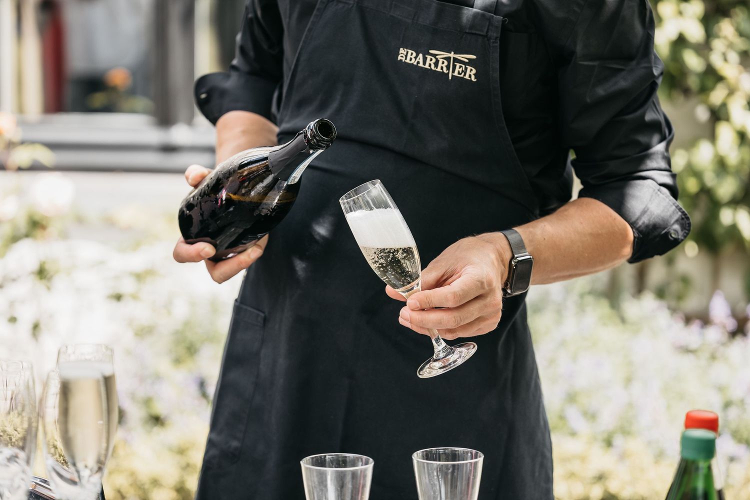 Wedding server in black apron pouring champagne into a flute glass at an outdoor reception.