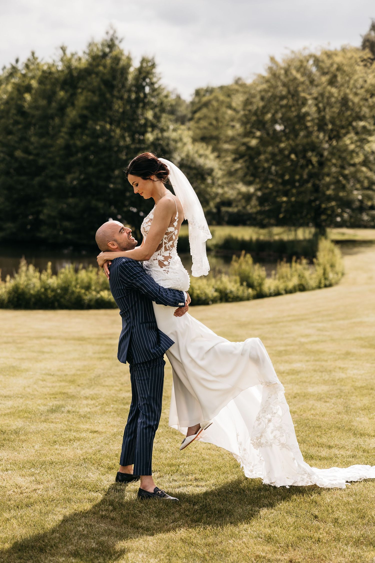 Groom in navy pinstripe suit lifts bride in lace wedding gown on sunny green lawn.