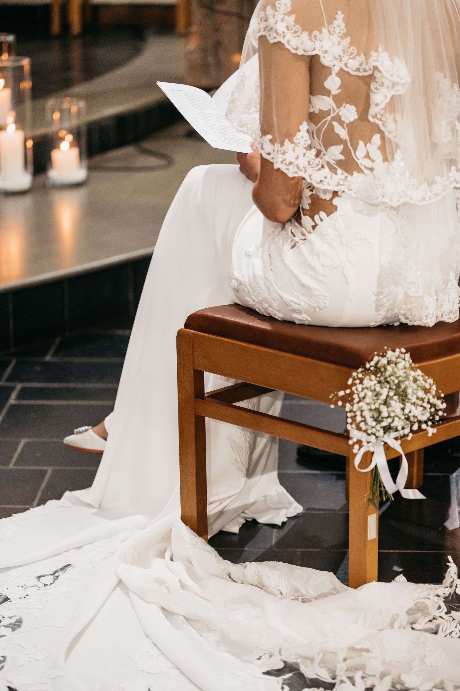 Bride in lace gown holds ceremony booklet with baby's breath bouquet at feet.
