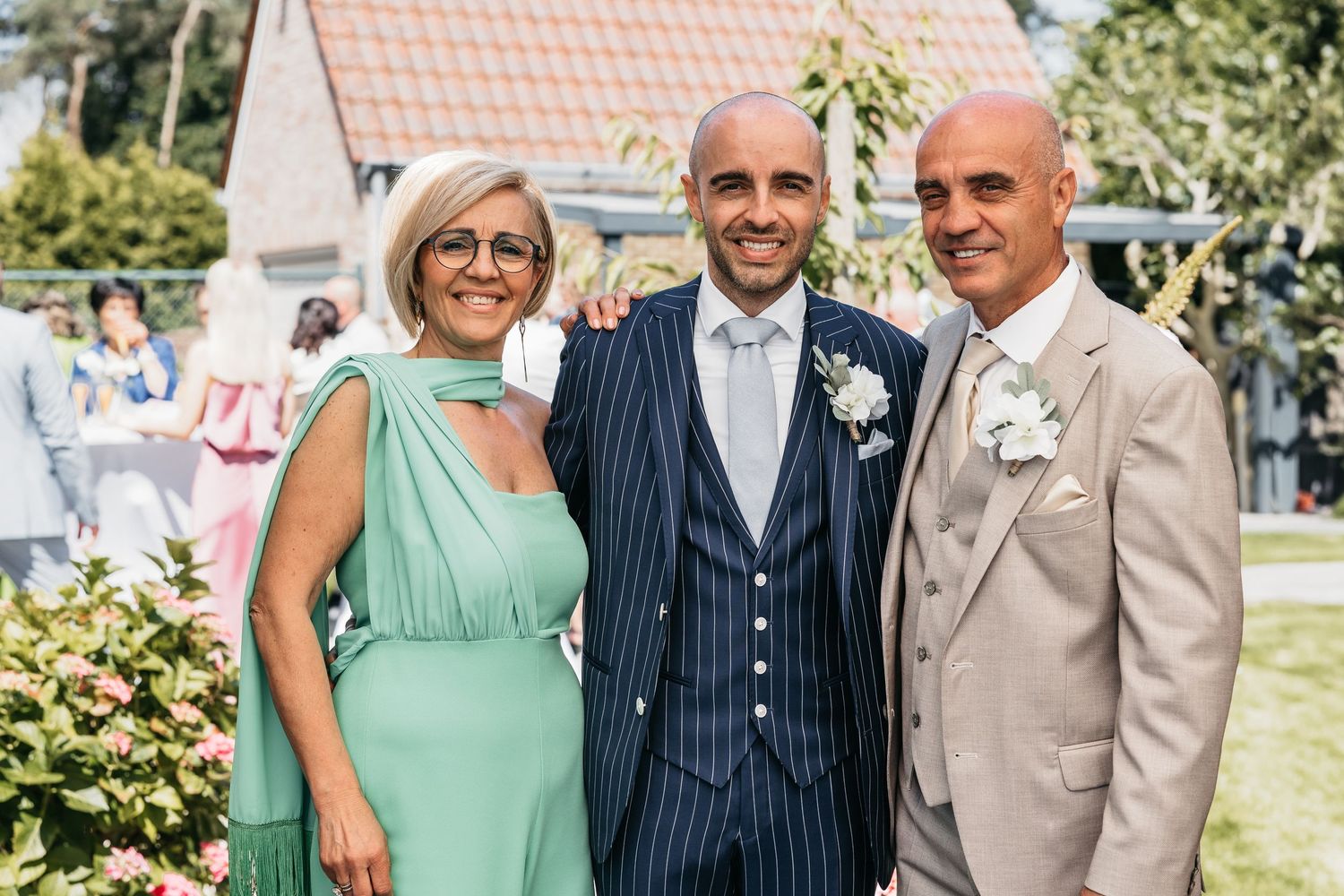 Happy groom in navy pinstripe suit smiling with elegantly dressed parents at outdoor wedding.