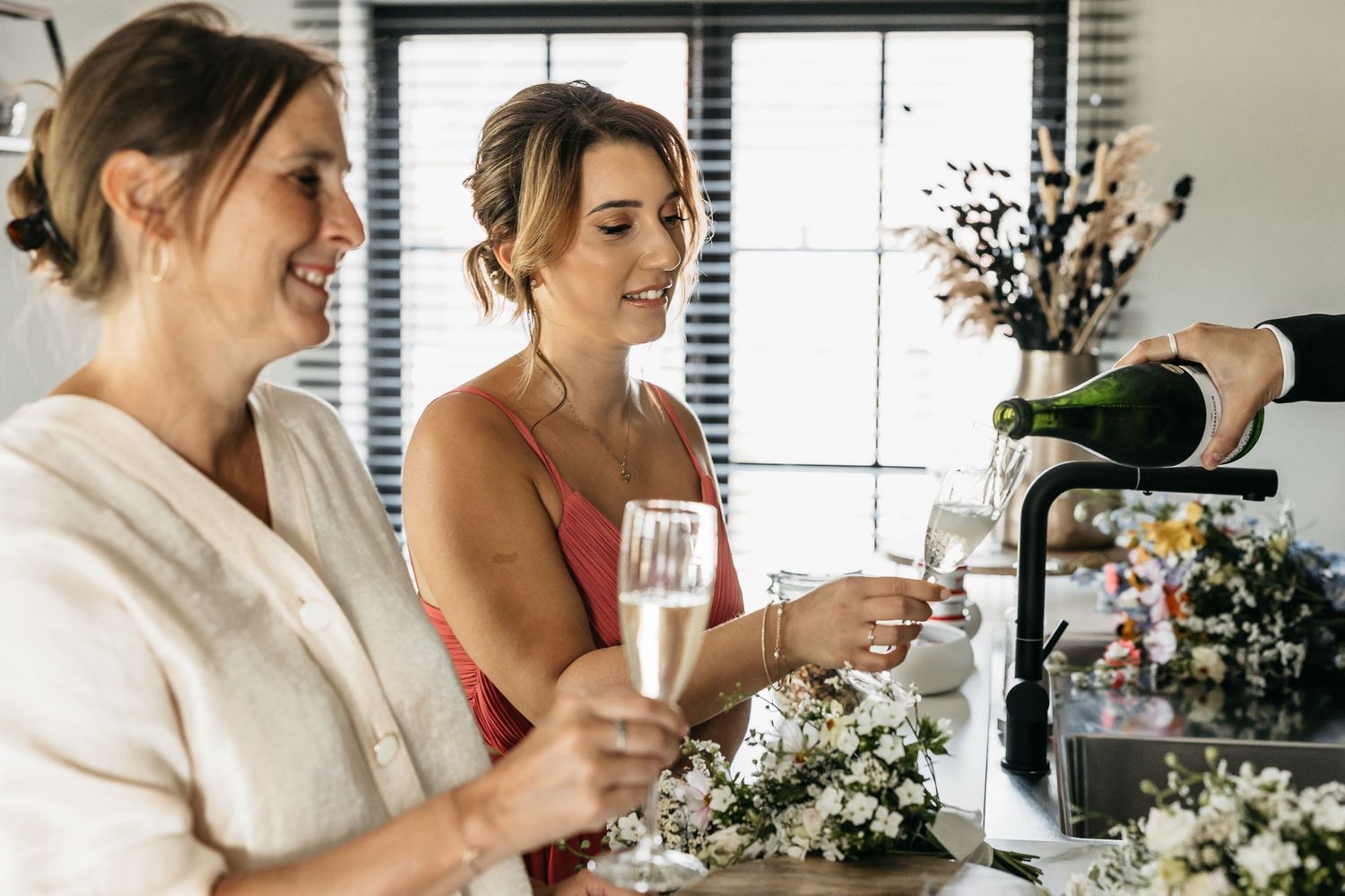 Two bridesmaids smiling while champagne is poured into flutes surrounded by white floral arrangements.