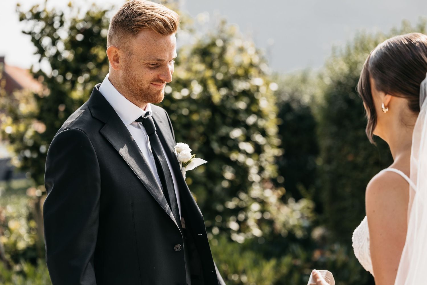 Groom in black suit smiles down shyly at bride during romantic outdoor wedding portrait in garden.