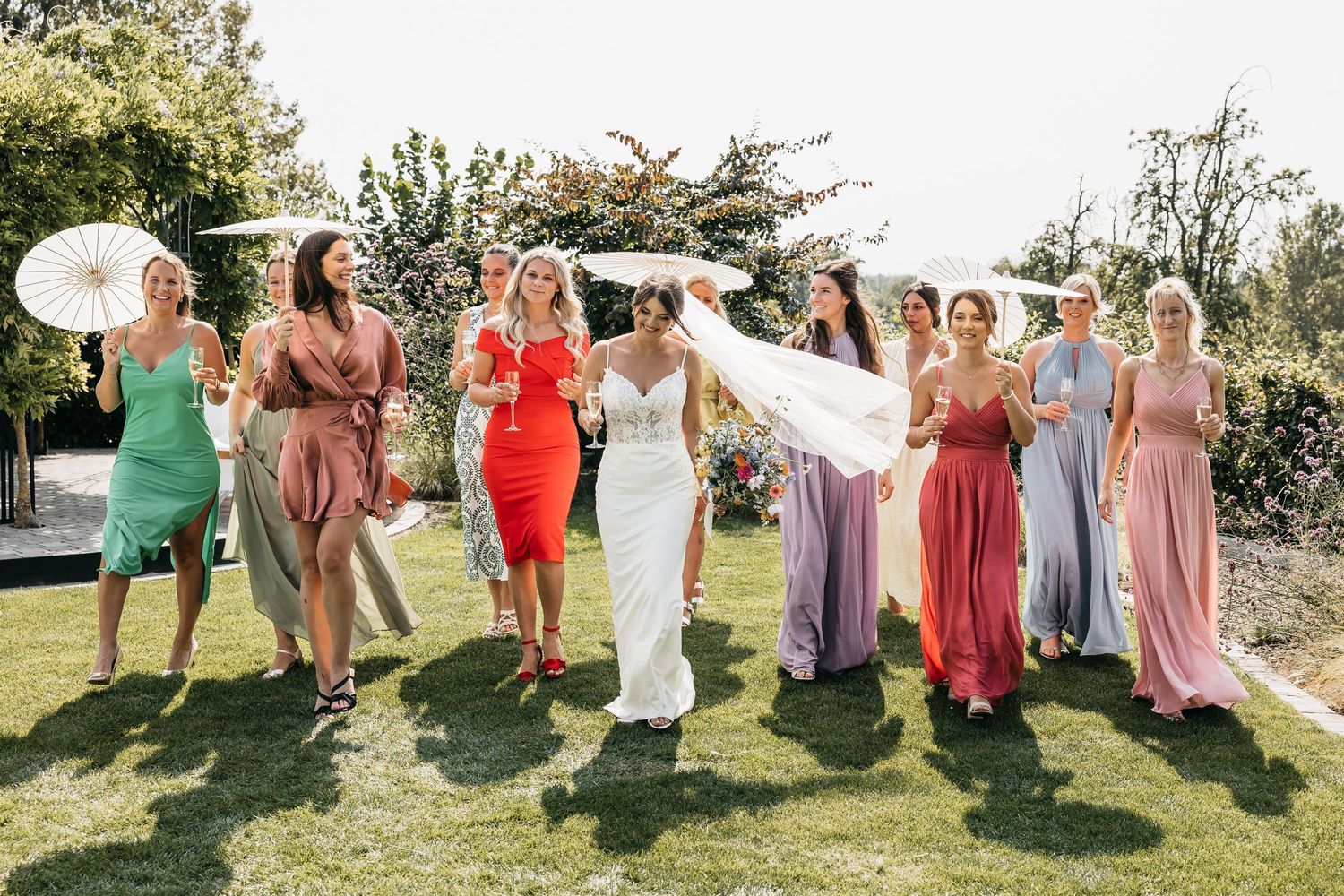 Bride lifting her dress while walking with bridesmaids in rainbow-colored gowns at sunny garden wedding.