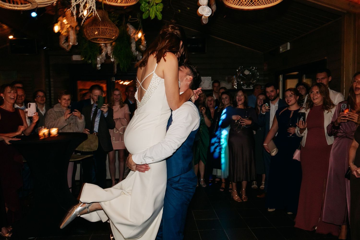Groom lifts bride joyfully during first dance at wedding reception surrounded by cheering guests in dimly lit venue.