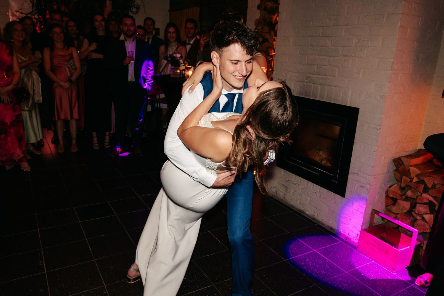 Groom dipping bride during energetic wedding reception dance near fireplace with purple lighting.