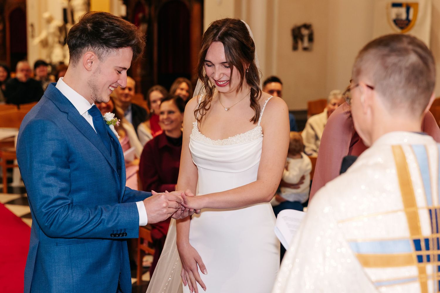 Groom in blue suit places wedding ring on brides finger during church ceremony.