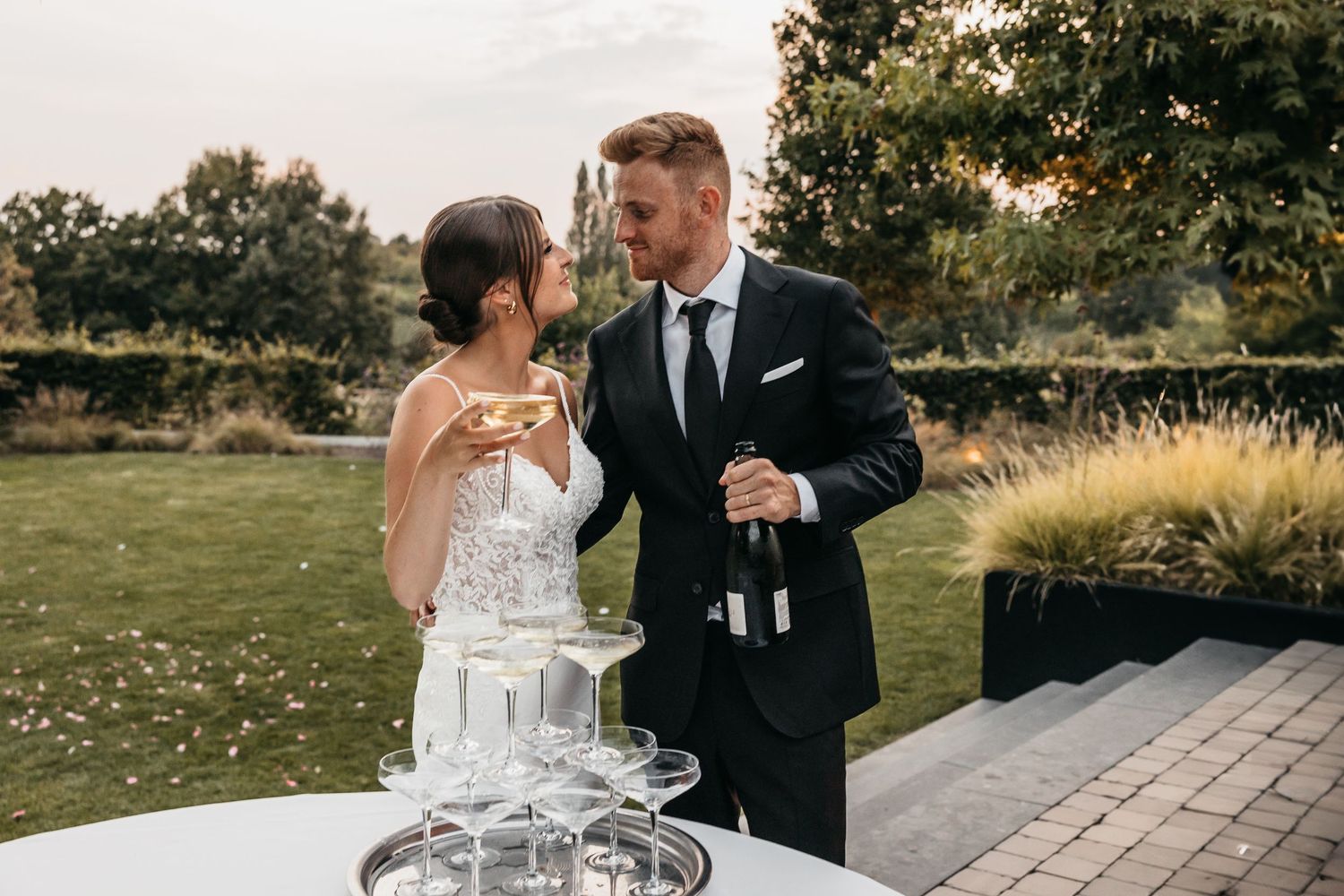 Bride and groom pouring champagne tower at outdoor wedding reception in garden setting.