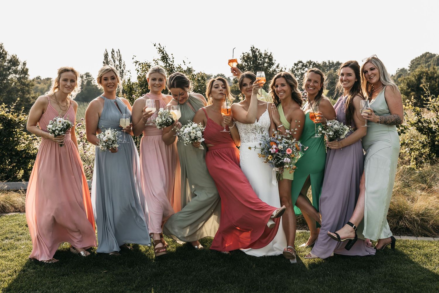 Bride and bridesmaids in multicolored dresses laugh and celebrate outdoors at a wedding.
