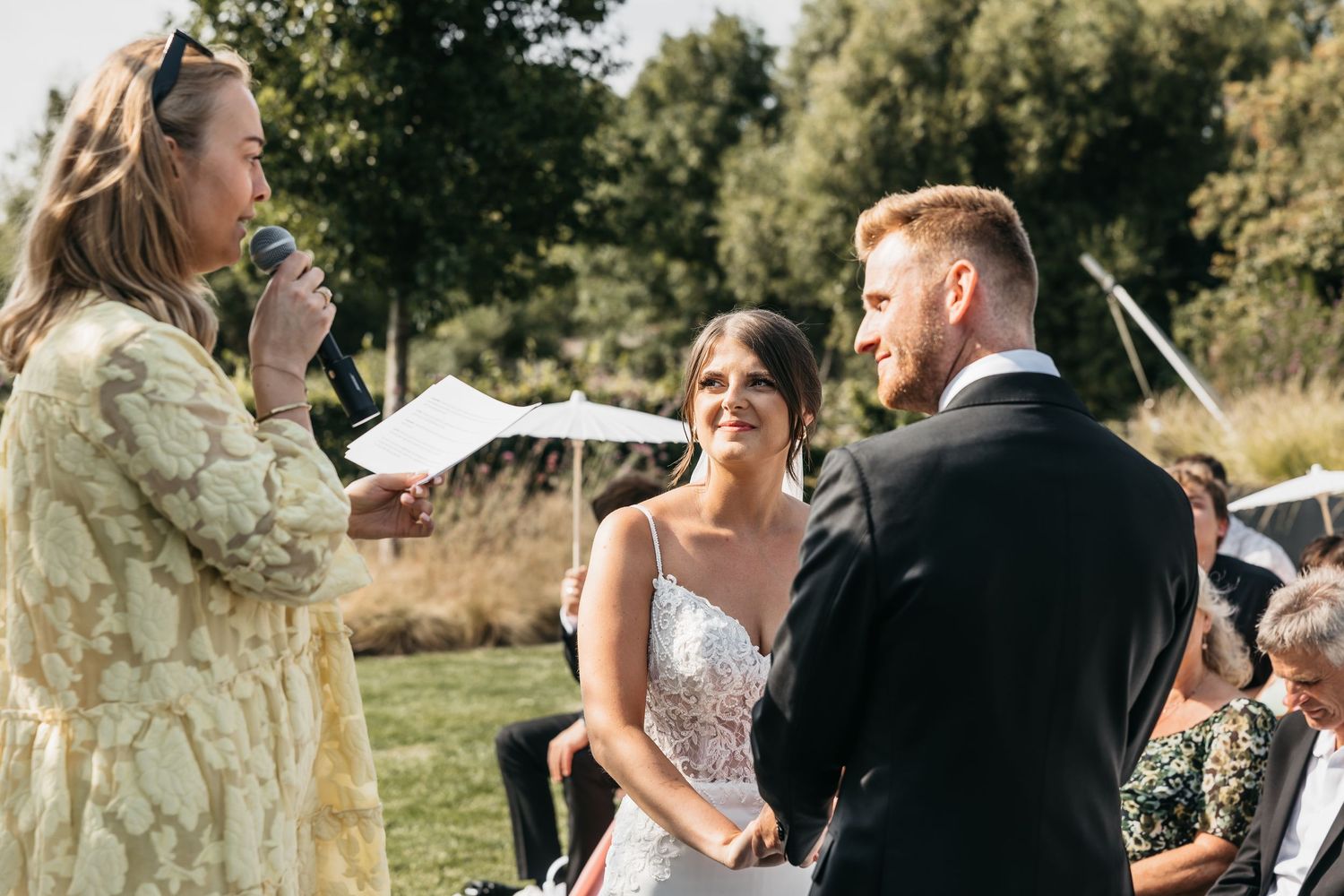 Officiant reads vows as smiling bride and groom face each other at outdoor wedding ceremony.