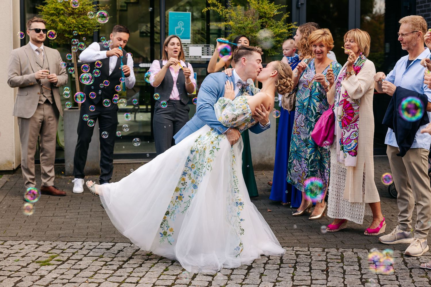 Newlyweds share a romantic dip kiss surrounded by guests blowing colorful soap bubbles.