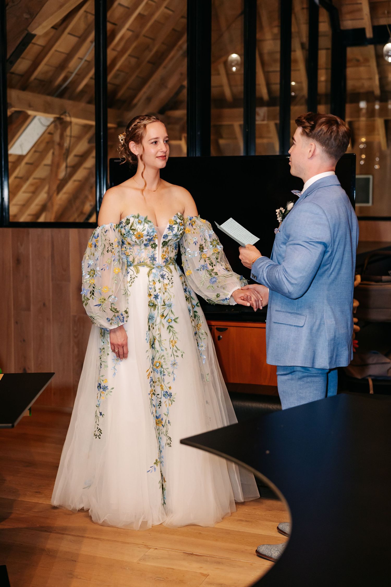 Bride in a floral embroidered gown holds hands with groom in a blue suit during wedding vows.