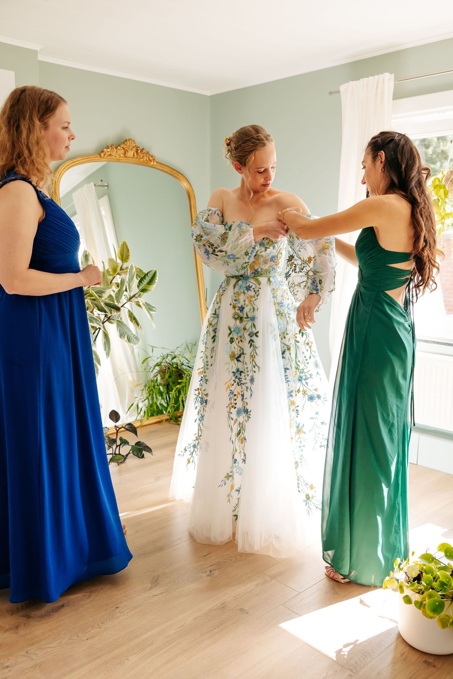 Two bridesmaids in blue and green gowns helping bride adjust floral wedding dress details.