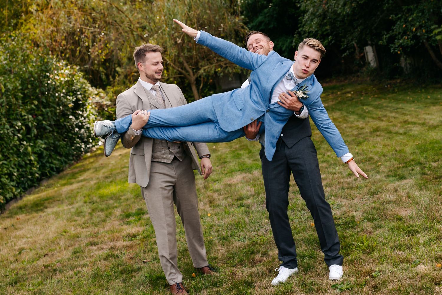 Three men in suits playfully pose with one being carried like an airplane in a garden setting.