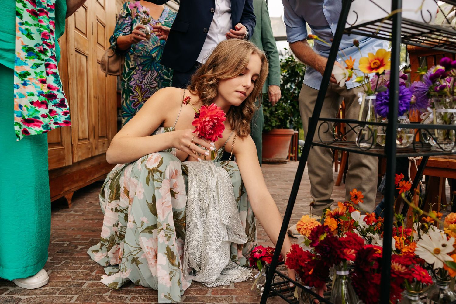 Woman in floral dress crouching to smell red dahlia at outdoor flower display.