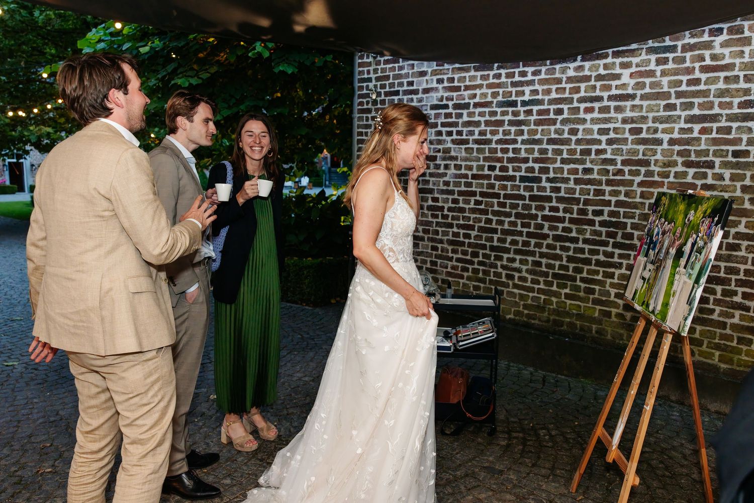 Bride in white gown admiring live wedding painting on easel at evening reception.