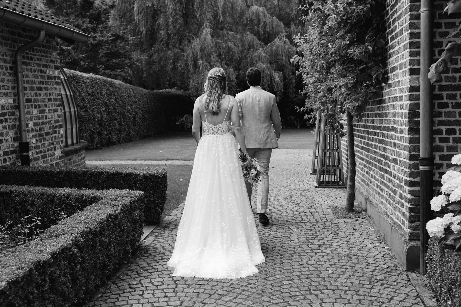 Black and white photo of bride and groom walking hand in hand along a cobblestone garden path.